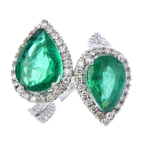An emerald and diamond crossover ring. Designed as two graduated pear-shape emeralds, each within a
