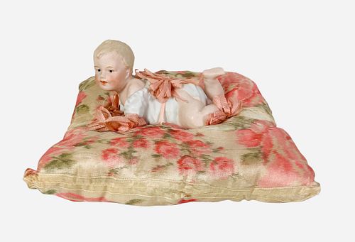 Gebruder Heubach Piano Baby. 5" long overall, bisque figure with molded and painted hair and facial features, intaglio eyes, tied to a silk pillow wit