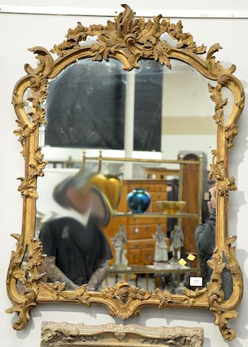A Louis XV/XVI Transitional Carved Giltwood Mirror, with a shaped top enclosed within a gilt wood frame with a molded border and carved with C-scrolls