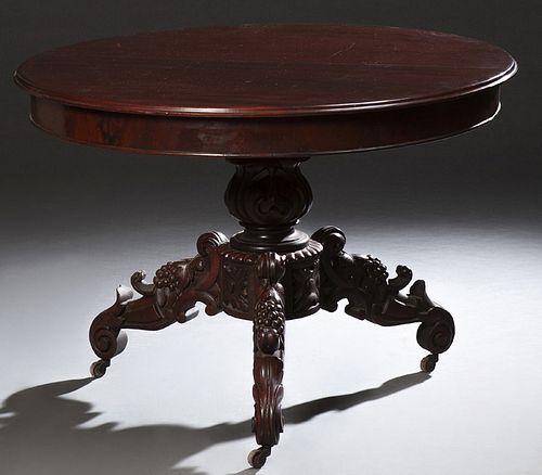 American Carved Mahogany Center Table, early 20th c., the ogee edge circular top on an urn form support to tripodal grape carved scrolled legs on cast