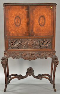 Carved walnut victrola cabinet having two burl walnut doors over elaborate carved figural drawer with base carved with horse carriag...