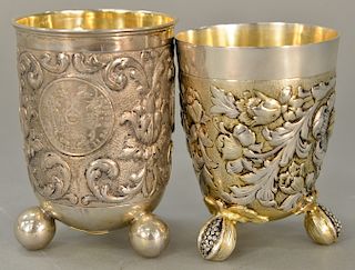 Two German silver beakers, each on three feet, one mounted with three large silver coins, one with floral chased body with gilt high...