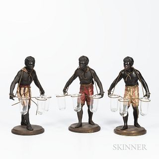 Three Blackamoor Table Figures, 19th century, each painted metal with variously painted breeches, and supporting three glass cups, ht.