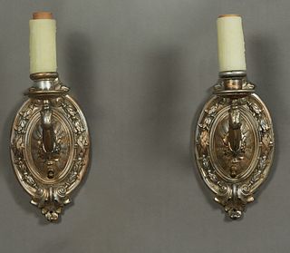 Pair of American Spelter Single Light Wall Sconces, c. 1930, the oval relief decorated back plate issuing a curved arm with a faux candle socket, H.- 