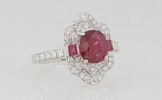 Lady's Platinum Dinner Ring, with a central oval ruby flanked by two ruby baguettes, atop a pierced shaped border of round diamonds, the shoulders of 