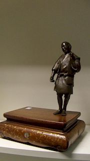 A late 19th/early 20th century bronze figure of a man standing to one corner of a wooden stand gold