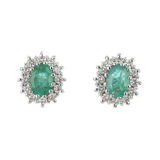 A pair of emerald and diamond cluster ear studs. Each designed as an oval-shape emerald, within a br