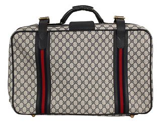 The suitcase has leather straps, piping, and handles, navy and red bands and gold hardware. The canvas suitcase is decorated with the Gucci monogram. 