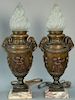 Pair of bronze torchere form table lamps with frosted flame form shades over bronze urn body with ram's head handles and mold