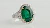 Lady's 18K White Gold Dinner Ring, with an oval 9.25 carat emerald atop a border of round diamonds, the tapered pierced shoulders with central graduat