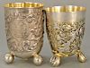 Two German silver beakers, each on three feet, one mounted with three large silver coins, one with floral chased body with gilt high...